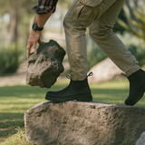 Person in Composite Toe Black Rainier Boots and cargo pants stepping onto a rock while lifting a large stone in an outdoor setting.