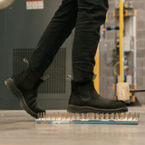 Person wearing Composite Toe Black Rainier Boots. Standing on a bed of nails in an industrial setting, demonstrating puncture resistance.