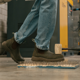Person wearing Composite Toe Rainier Boots Olive Green. Standing on a bed of nails in an industrial setting, demonstrating puncture resistance.