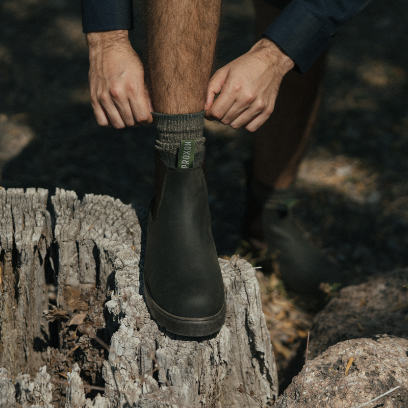 Close-up of a person adjusting their sock while wearing Soft Toe Rainier Boot Black, standing on a tree stump outdoors.