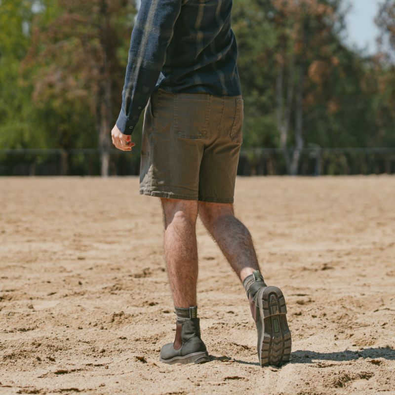 Man walking on land wearing the Soft Toe Rainier Boots Olive Green, shorts, and a long-sleeve shirt in an outdoor setting.