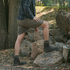 Man wearing black Soft Toe Rainier Boots and shorts, stepping on rocks in a wooded outdoor setting