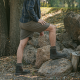 Man wearing black Soft Toe Rainier Boots and shorts, stepping on rocks in a wooded outdoor setting