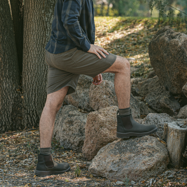 Man wearing black Soft Toe Rainier Boots and shorts, stepping on rocks in a wooded outdoor setting