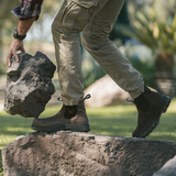 Person in Composite Toe Rainier Boots Tobacco and cargo pants stepping onto a rock while lifting a large stone in an outdoor setting.