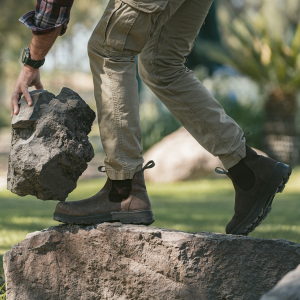 Person in Composite Toe Rainier Boots Tobacco and cargo pants stepping onto a rock while lifting a large stone in an outdoor setting.