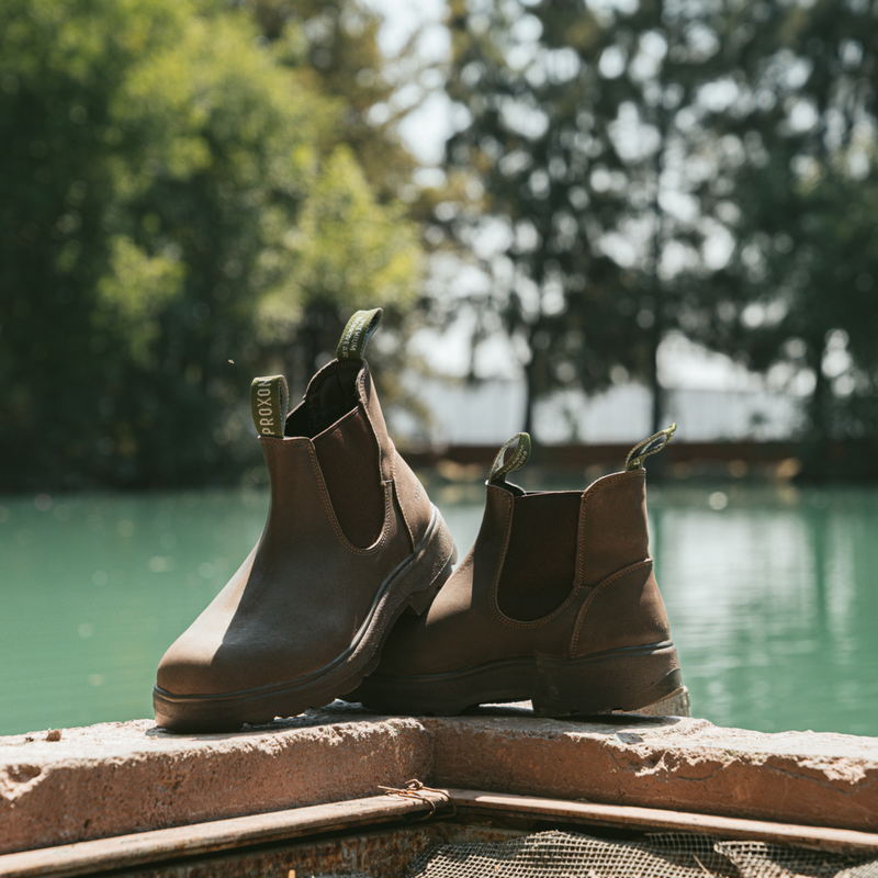 Pair of Soft Toe Rainier Boots Tobacco displayed on a ledge by a lake, with trees in the background.