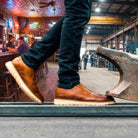 Person wearing brown leather shoes stepping onto a metal platform with a bar and industrial setting in the background.
