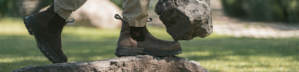 Collection Banner. Person in Composite Toe Black Rainier Boots and cargo pants stepping onto a rock while lifting a large stone in an outdoor setting. 