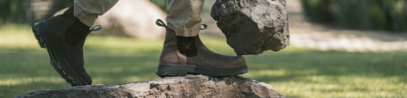 Collection Banner. Person in Composite Toe Black Rainier Boots and cargo pants stepping onto a rock while lifting a large stone in an outdoor setting. 